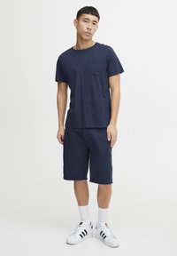 Námořnická modrá trička s krátkým rukávem s přední kapsou, v kombinaci s námořnickými šortkami do délky ke kolenům a bílými teniskami s černými pruhy.