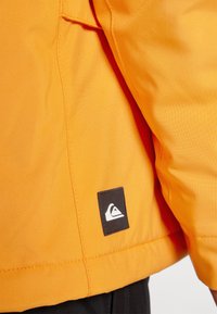 Oranžis jakikäte varrukas tekstuuriga kangast musta logoga plaastriga, millel on valge mäe disain, esitleb kontrastseid värve ja puhtaid jooni.