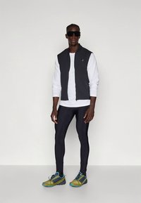 Gilet nero senza maniche sopra una camicia bianca a maniche lunghe, abbinato a leggings neri e scarpe da ginnastica verdi con dettagli blu.