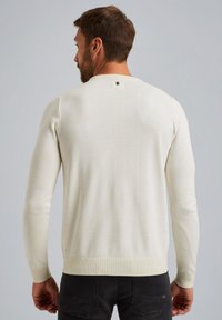 Maglione beige chiaro con maniche lunghe, polsini e orlo a costine. Presenta un piccolo tag di branding all'collo. Tessuto lavorato a maglia liscio.