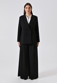 Frau steht mit dem Gesicht nach vorne, trägt ein schwarzes doppelt knöpfbares Blazer und weit geschnittene schwarze Hosen über einer weißen Bluse mit einem silbernen Verschluss.