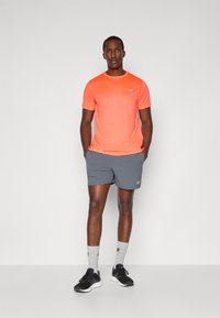 Lys oransje trenings T-skjorte med kortermede, kombinert med grå shorts. Svarte joggesko med grå ankelstrømper. Enkel, sporty design.