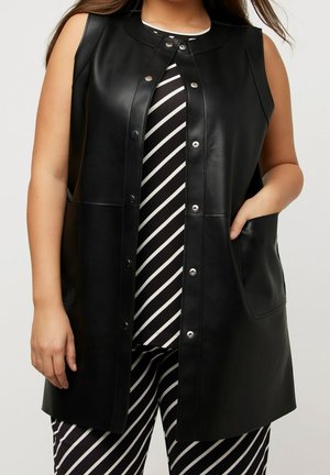 Femme portant un gilet en cuir noir sans manches boutonné sur une tenue rayée diagonale noire et blanche, une main dans la poche du gilet.