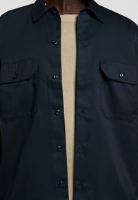 Camicia azzurra navy a maniche lunghe realizzata in tessuto liscio, con due tasche frontali e una t-shirt beige visibile sotto.