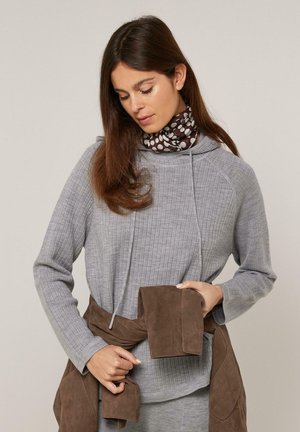 Frau mit langen braunen Haaren, die einen grauen gerippten Pullover und einen gepunkteten Schal trägt, hält eine braune Jacke, die um ihre Taille gebunden ist.