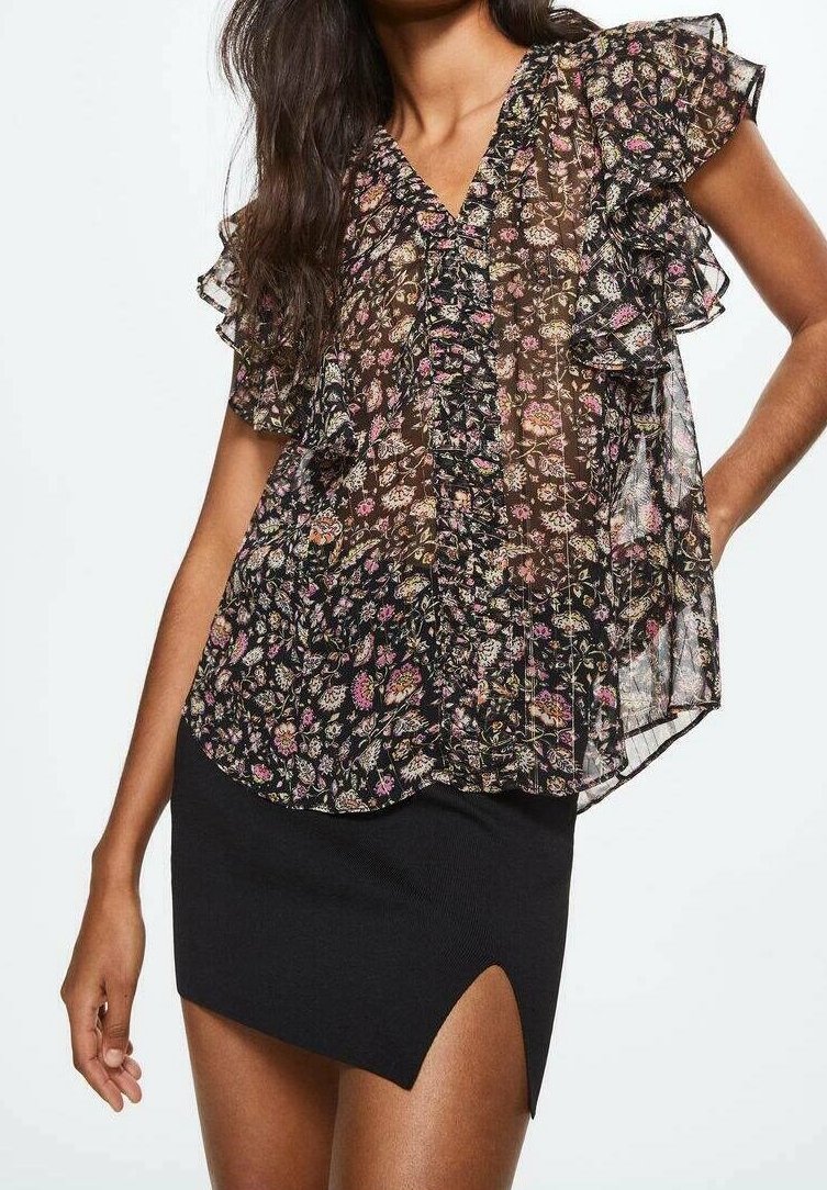 Femme portant un chemisier noir transparent à motifs floraux avec des manches à volants et une mini-jupe noire avec une fente devant, posant devant un fond uni.