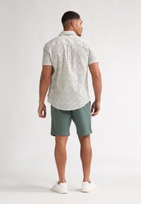 Cămașă cu mâneci scurte și imprimeu floral în alb, cu modele albastre și verzi, combinată cu pantaloni scurți verzi. Include tiv curbat și detaliu la spatele yoke-ului.