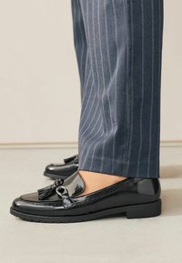 Patentierte schwarze Loafer mit Quasten und Hardware-Akzenten, getragen mit navyblauen Nadelstreifenhosen, die ein schlankes, poliertes Design zeigen.