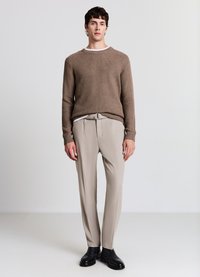 Brauner, strukturierter Strickpullover, beige maßgeschneiderte Hose mit vorderen Falten, schwarze glatte Schuhe und eine minimalistische silberne Gürtelschnalle.
