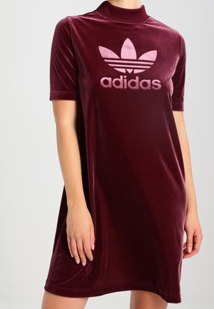 Woman wearing a short-sleeve burgundy velvet Adidas dress with a high collar and logo on the chest against a plain background.