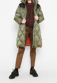 Cappotto lungo imbottito trapuntato in verde oliva con accenti neri sulle spalle e foderato in arancione, abbinato a pantaloni marroni e scarpe nere stringate.
