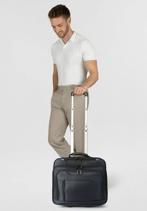 Joven sonriente, de pie y sujetando el mango extendido de una maleta negra con ruedas y bolsillos frontales.