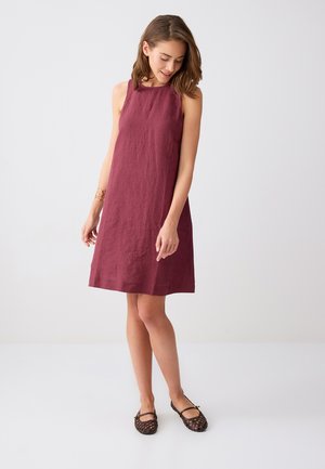 Donna che indossa un abito senza maniche al ginocchio di colore rosa scuro e ballerine intrecciate scure, guarda verso il basso con le mani rilassate lungo i fianchi, sfondo neutro.