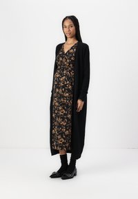 Cardigan lungo nero sopra un vestito floreale nero con accenti beige. Il vestito presenta una scollatura a V e una vita con fiocco. Le scarpe con tacco nere completano il look.