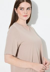 Beige V-kaula-aukkoinen T-paita, valmistettu pehmeästä kankaasta, jossa on lyhyet hihat ja rento istuvuus, hienovaraisella saumaonnilla olkapäiden kohdalla.