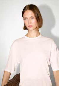 Lichtroze katoenen t-shirt met een ronde halslijn en korte mouwen. Model houdt een geweven bruine tas vast, met zachte verlichting die de texturen accentueert.