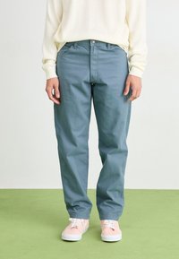 Light blue cotton chinos with a relaxed fit, front pockets, and a button closure, paired with light pink sneakers on a green background.