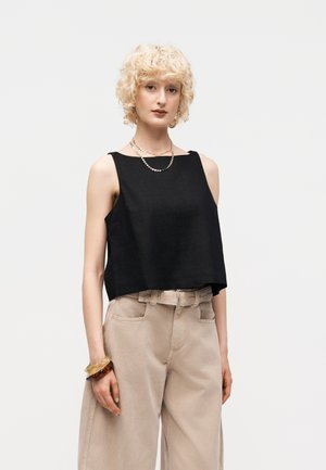 Νεαρό άτομο με κοντά σγουρά ξανθά μαλλιά, που φοράει μαύρο αμάνικο τοπ, μπεζ παντελόνι με φαρδιά πόδια και ψηλή μέση, κολιέ και βραχιόλια.