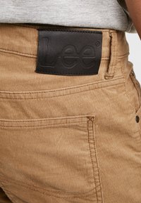 Béžové kordové kalhoty mají texturovaný povrch a na zadním pásku černý kožený štítek s embosovaným nápisem "Lee".