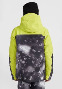 Helle gelbe Skijacke mit einem schwarz-grauen, raumthemenartigen Muster. Mit Kapuze, kontrastierenden Ärmeln und auffälligem Logodetail.