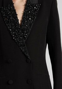 Blazer nero con risvolto in paillettes ornato di lucenti perline nere, design doppiopetto e bottoni neri rotondi.