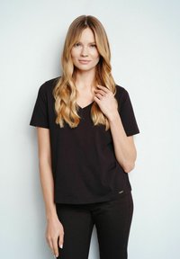 Czarna koszulka polo z dekoltem w serek, wykonana z miękkiego materiału, z krótkimi rękawami i luźnym krojem. Zauważalna metka marki przy dolnym brzegu.