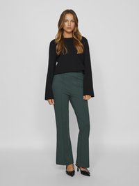 Haut noir à manches longues associé à un pantalon évasé vert foncé. Tissu texturé avec une finition lisse. Des escarpins noirs à bout pointu complètent la tenue.