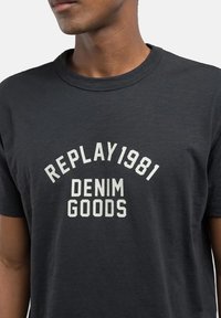 Camiseta de algodón gris oscuro con texto impreso en blanco: "REPLAY 1981" en un arco sobre "DENIM GOODS" en un estilo en negrita y de bloques. Cuello redondo clásico.