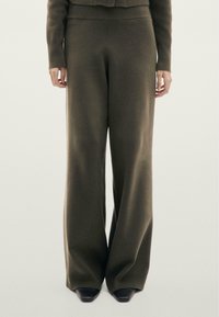 Personne portant un pantalon large en maille vert olive et un pull assorti à manches longues, debout devant un fond clair uni.