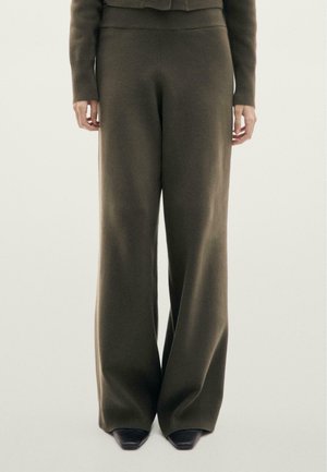 Osoba stojící proti jednobarevnému světlému pozadí, oblečená v olivově zelených širokých pletených kalhotách a sladěném svetru s dlouhými rukávy.