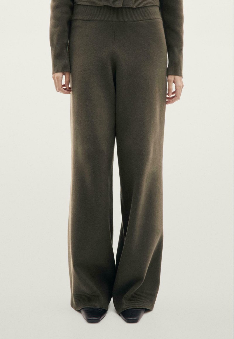 Personne portant un pantalon large en maille vert olive et un pull assorti à manches longues, debout devant un fond clair uni.