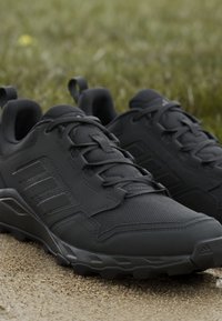 Czarne buty sportowe z siateczkowym wierzchem, teksturowanymi akcentami i gumowymi podeszwami. Funkcje obejmują wzmocniony nosek i płaskie sznurowadła.