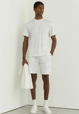 Mladý muž v bielych krátkych šortkách s textúrou a krátkym rukávom košele, drží zladenú bundu, stojí pri bielej stene.