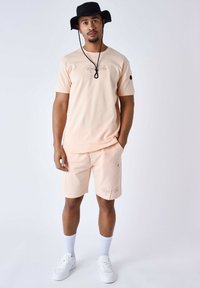 T-shirt rosa chiaro con la scritta "Project Paris", abbinata a pantaloni corti coordinati. Sneakers e calzini bianchi, e un cappello a secchiello nero completano il look.