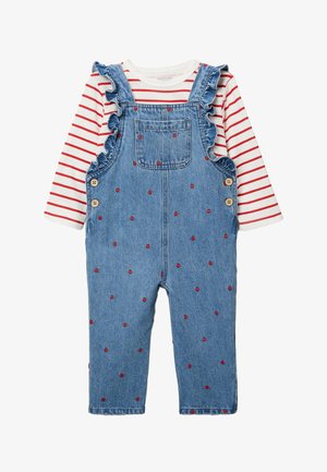 Salopette en denim avec de petits motifs de cœurs rouges et des bretelles volantées, portée sur un chemisier blanc à manches longues avec des rayures horizontales rouges.