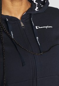 Felpa nera con zip realizzata in tessuto morbido, dotata di zip frontale e logo bianco sul petto, con corde di regolazione a motivi all'interno del cappuccio.
