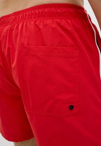 Bañador rojo hecho de material ligero, con una cinturilla elástica, franjas laterales y un bolsillo trasero con un detalle de ojete negro.