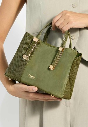 Main tenant un petit sac à main vert olive avec des accents dorés, devant en daim, côtés texturés, et logo "Dune London", sur un vêtement beige.