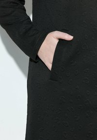 Abrigo negro texturizado con un bolsillo y un sutil patrón en relieve. La tela parece resistente, con un diseño estructurado que realza la silueta.