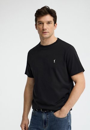 Man with dark hair wearing a black short-sleeve T-shirt with small white polo player logo and blue jeans, standing against white background.