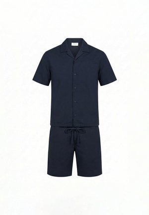 Ung mand, der står i profil iført en mørkeblå kortærmet skjorte med knapper, matchende shorts og sorte slip-on sko.