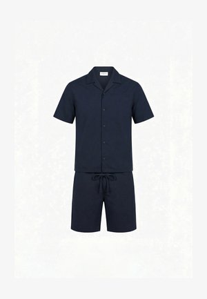 Ung mand, der står i profil iført en mørkeblå kortærmet skjorte med knapper, matchende shorts og sorte slip-on sko.