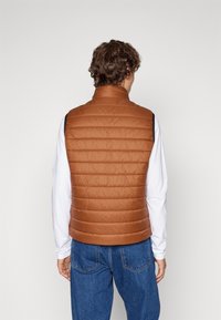 Braune, isolierte ärmellose Weste mit gestepptem Design, hohem Kragen und schwarzen Akzenten; getragen über einem weißen Langarmshirt und blauen Jeans.