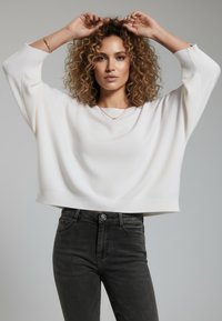 Weißer, gekürzter Pullover mit weiten Ärmeln und geripptem Saum, kombiniert mit schwarzen Jeans. Das Model zeigt einen neutralen Gesichtsausdruck vor einfachem Hintergrund.