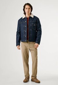 Τζιν μπουφάν με κρεμ γιακά από shearling, ύφασμα σκούρου μπλε και τσέπη στο στήθος. Συνδυασμένο με ανοιχτό μπεζ παντελόνια και καφέ αθλητικά παπούτσια.