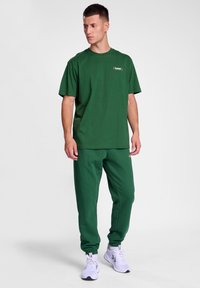 Zaļs kokvilnas T-krekls ar īsām piedurknēm un mazu logo krūtīs, kopā ar saskaņojošām zaļām sporta biksēm un baltām sporta apaviem.