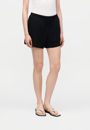 Kvinde iført sorte snor-shorts, hvid ærmeløs top og beige flade sandaler, der står foran en ensartet lys baggrund.