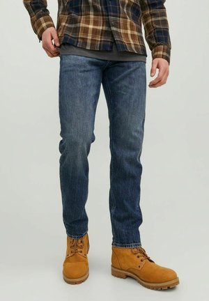 Man wearing blue jeans, brown plaid shirt, and tan lace-up boots standing against a plain light background.