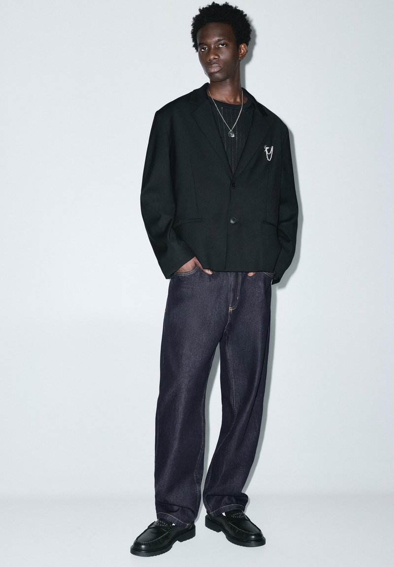Jeune homme portant un blazer noir avec une épingle chaîne, chemise en maille foncée, jean large foncé, chaussures noires, debout les mains dans les poches contre un mur blanc.