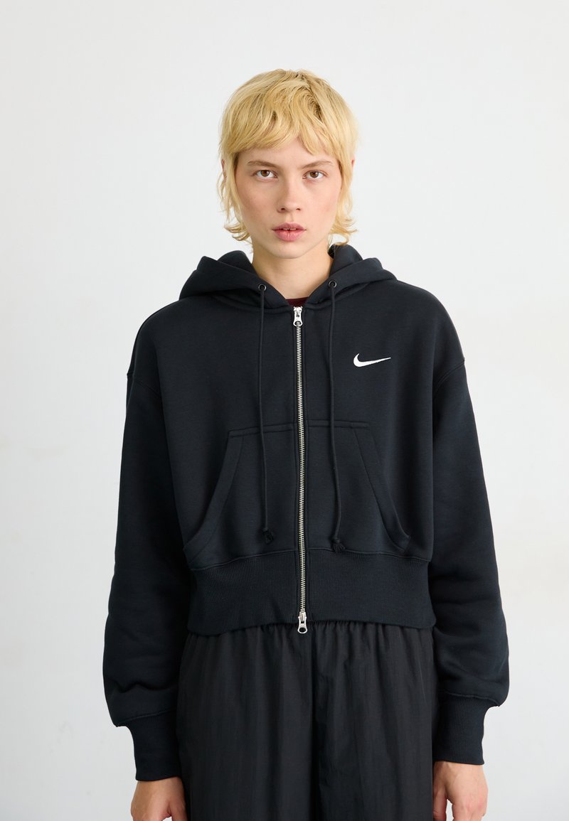 Haut de survêtement noir zippé avec un design court, fabriqué dans un matériau épais. Présente une poche avant, une capuche réglable avec cordon et un logo blanc.